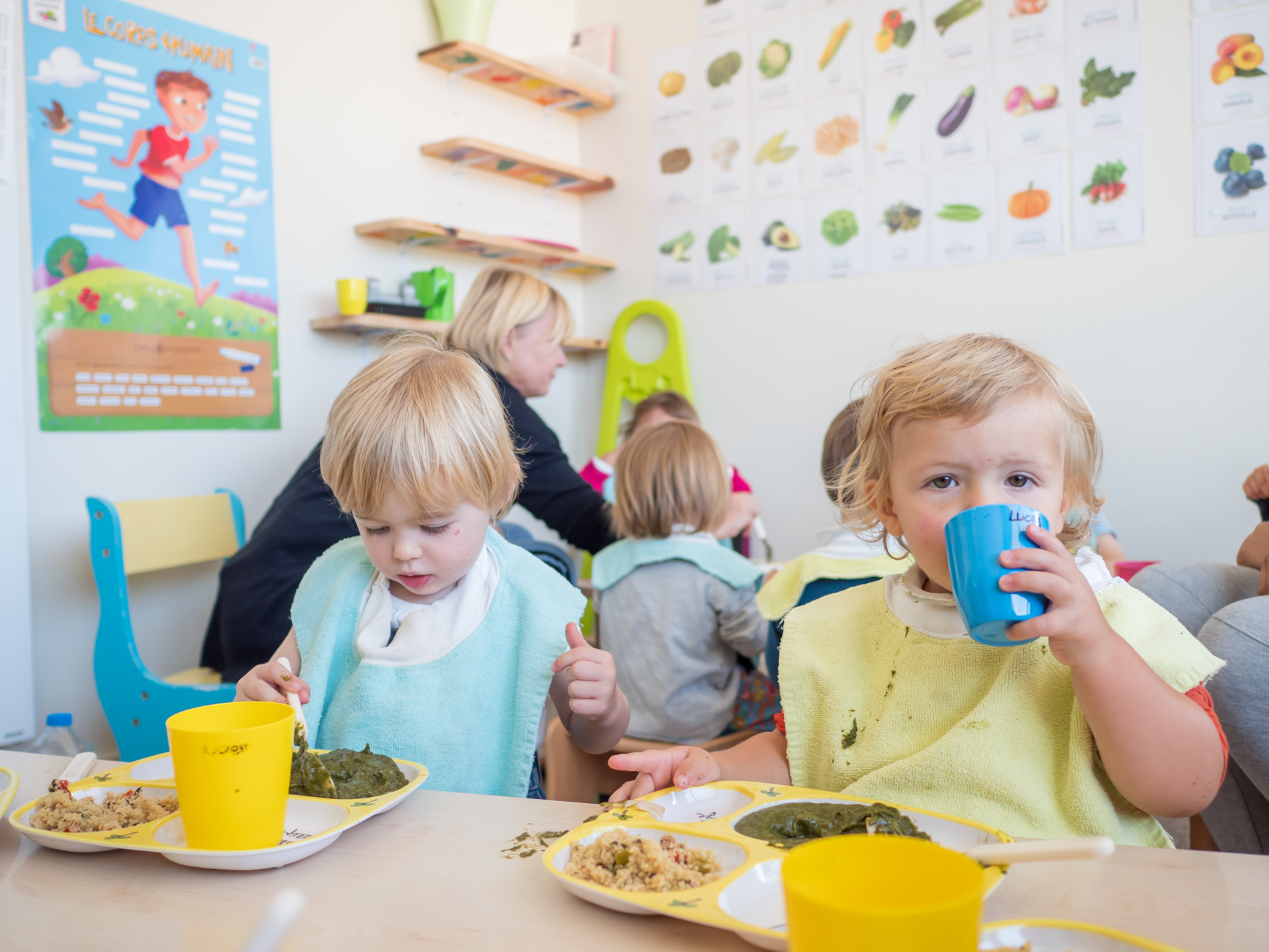 En premier plan, deux enfants mangent en autonomie, en arrière-plan les professionnelles en surveillance
