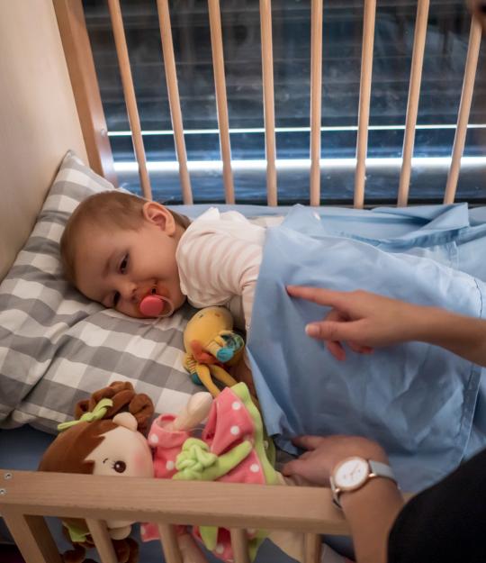 enfant dans son lit à barreau, avec une tétine, et une professionnelle qui pose la main sur lui