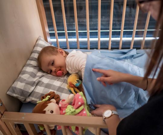 Nos professionnelles de la petite enfance accompagnent les enfants accueillis sur les temps de siestes. enfant dans son lit à barreau, avec une tétine, et une professionnelle qui pose la main sur lui