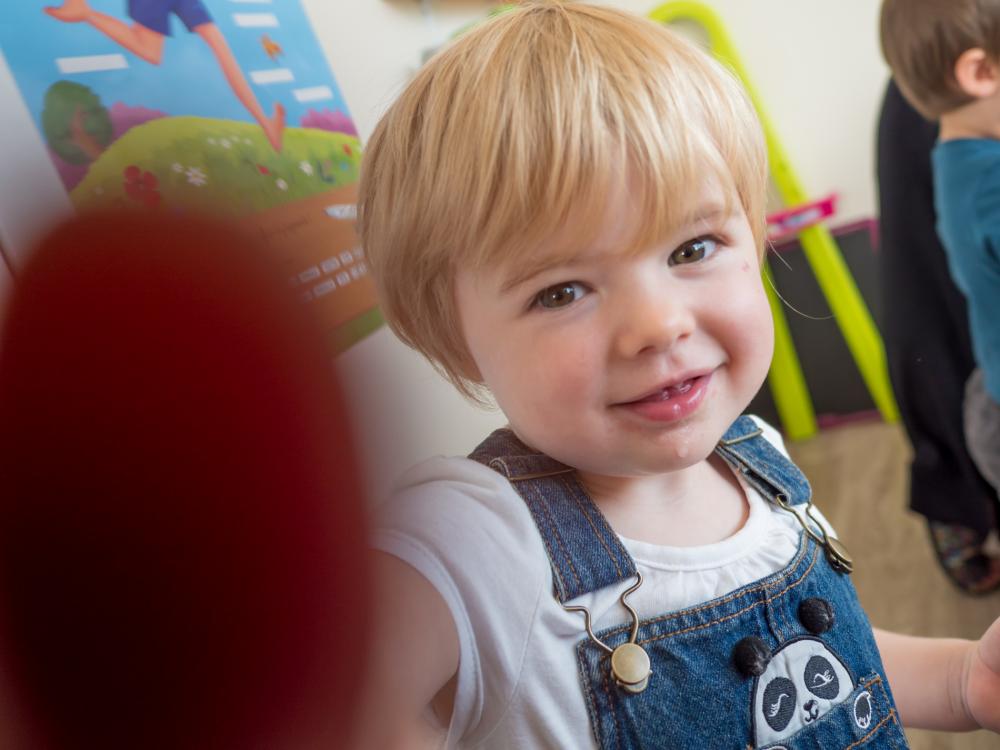Un jeune enfant blond en salopette bleu regarde l'appareil qui le prend en photo et le touche du doigt avec un sourire